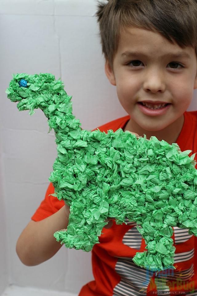 _ 2 Child holding a green, textured dinosaur craft made from paper, smiling in front of a white background, wearing a red shirt. | Sky Rye Design Child holding a green, textured dinosaur craft made from paper, smiling in front of a white background, wearing a red shirt.