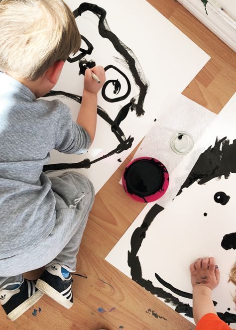 Child painting abstract art on floor with black ink, wearing gray outfit. Creative kids' activity in a home setting with wooden floor.