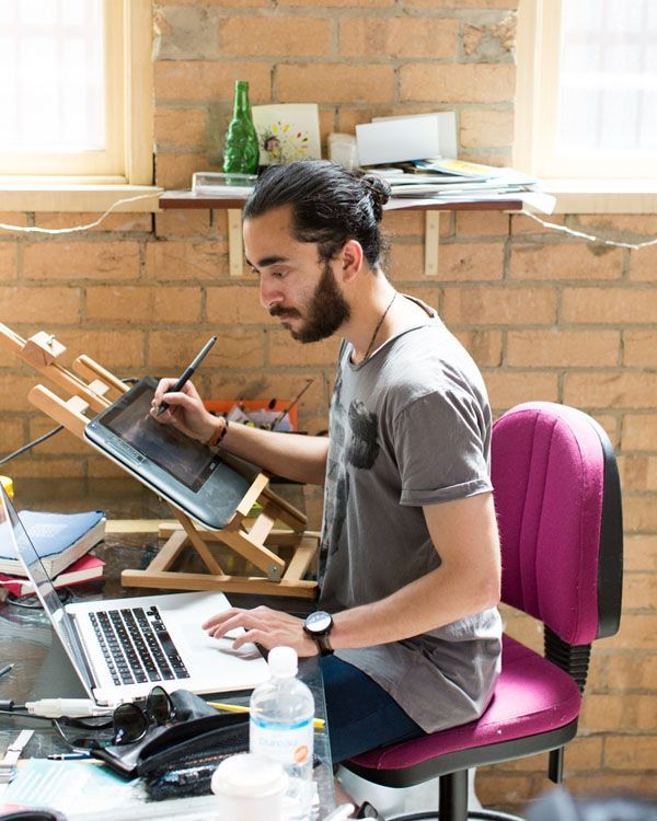Man working on digital art using a tablet and laptop in a creative home office with exposed brick walls.