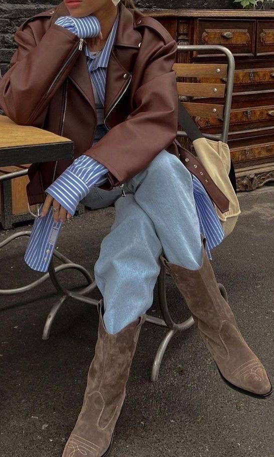 Person in brown leather jacket, striped shirt, light jeans, and suede boots sitting at a cafe table with wooden chairs. Fashionable look.