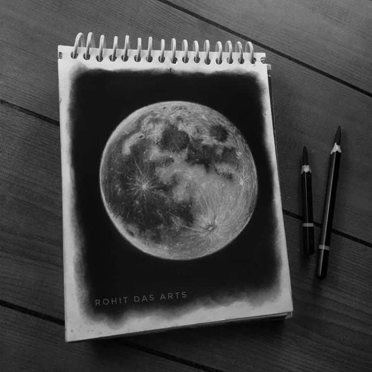 Sketch of a detailed moon on a spiral notebook with pencils on a wooden table.