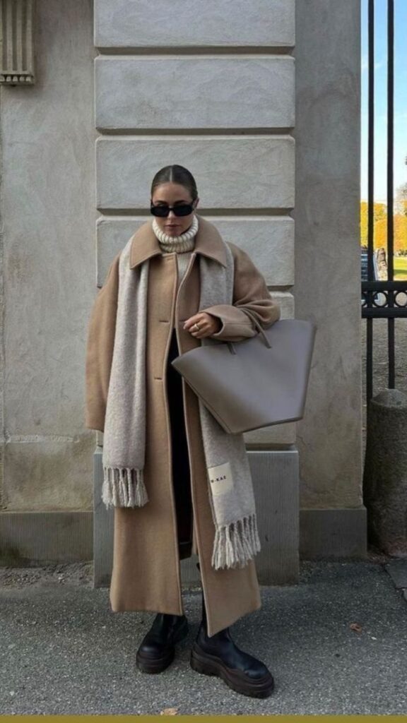 Stylish woman in a beige coat and scarf with oversized bag, sunglasses, and black boots, standing outdoors.