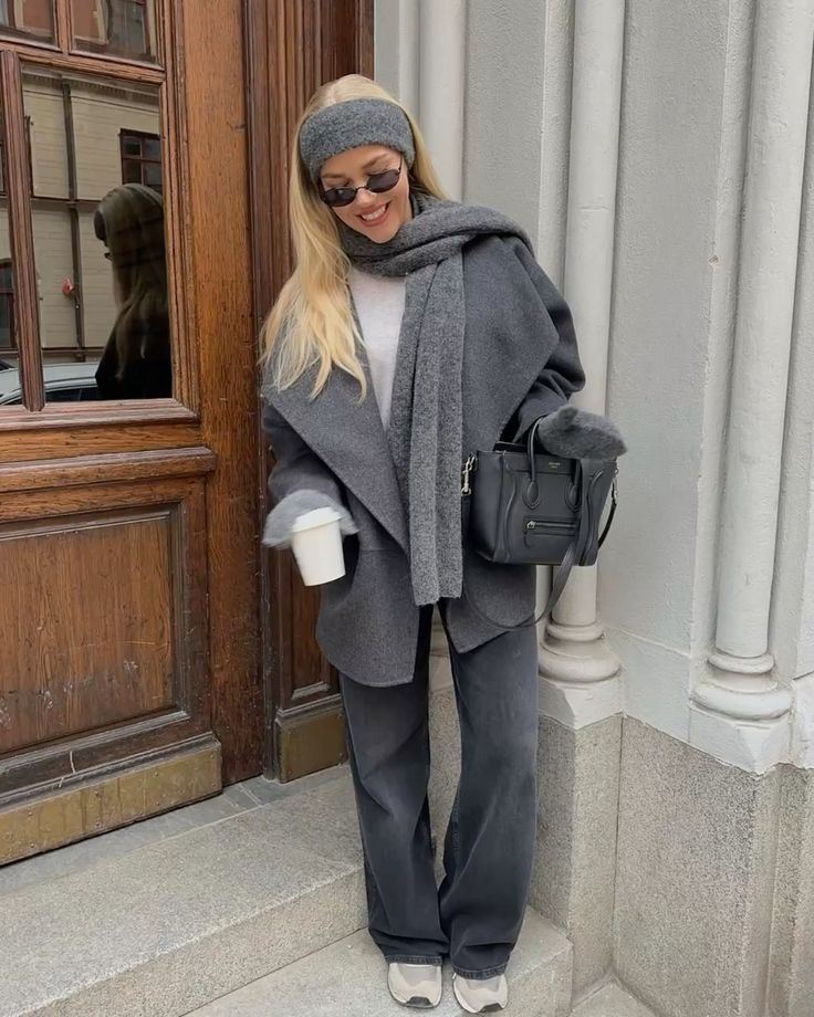 Fashionable woman in gray coat with scarf and headband holding coffee, standing outside a building in casual winter attire.