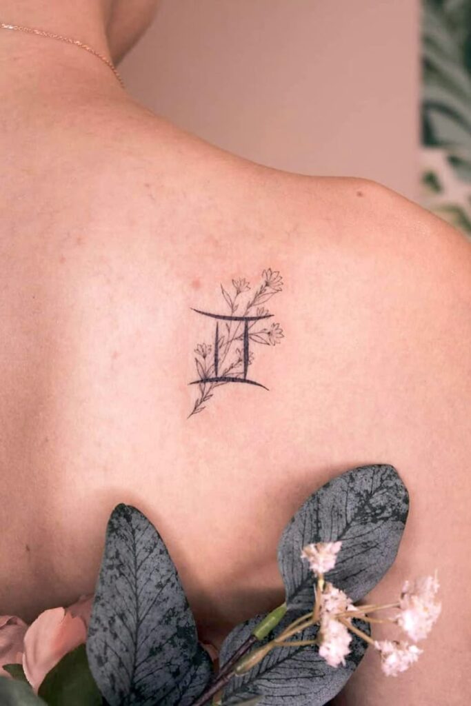 Gemini zodiac tattoo with floral design on shoulder, surrounded by delicate leaves and flowers.