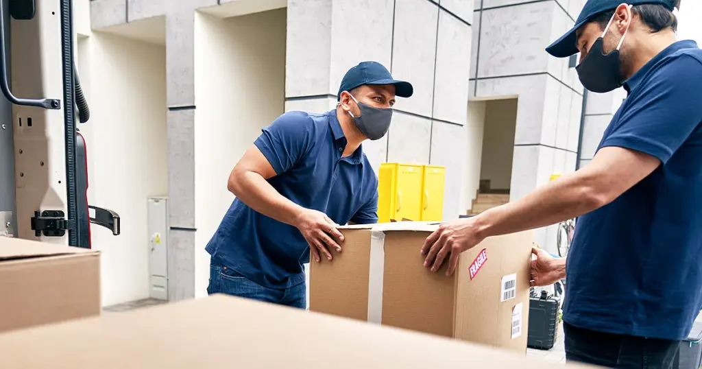 Two delivery workers in blue uniforms and masks handle a large cardboard box labeled fragile outside a building.