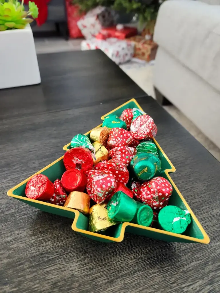 Festive chocolates in red, green, and gold wrappers fill a Christmas tree-shaped dish; holiday treats on a table.
