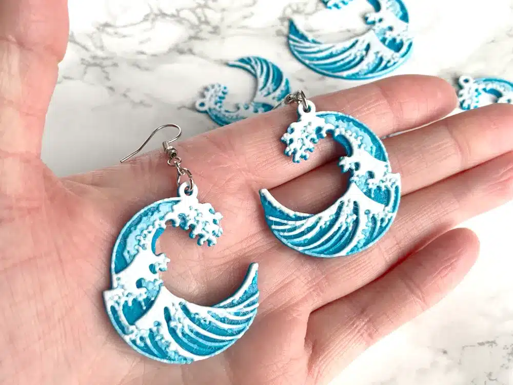 Hand holding blue wave-design earrings, inspired by ocean waves, with intricate details, lying on a marble surface.