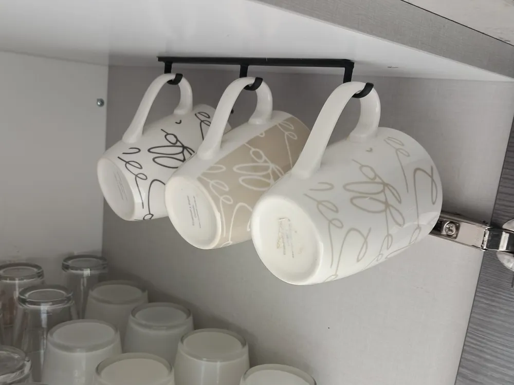 Three ceramic mugs hang under a cabinet beside stacked glasses in a kitchen, optimizing space with an efficient storage solution.
