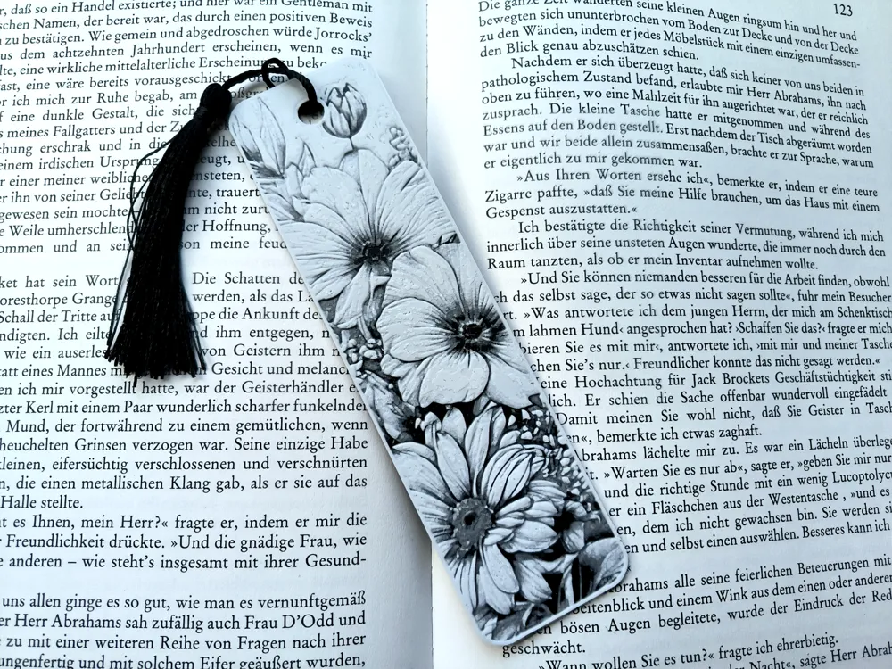 Floral bookmark with tassel resting on open German book, highlighting a passage. Perfect for book lovers and reading enthusiasts.