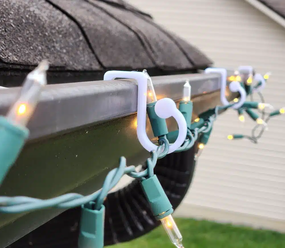 Close-up of holiday string lights attached to a roof gutter with white clips, glowing warmly against a residential background.