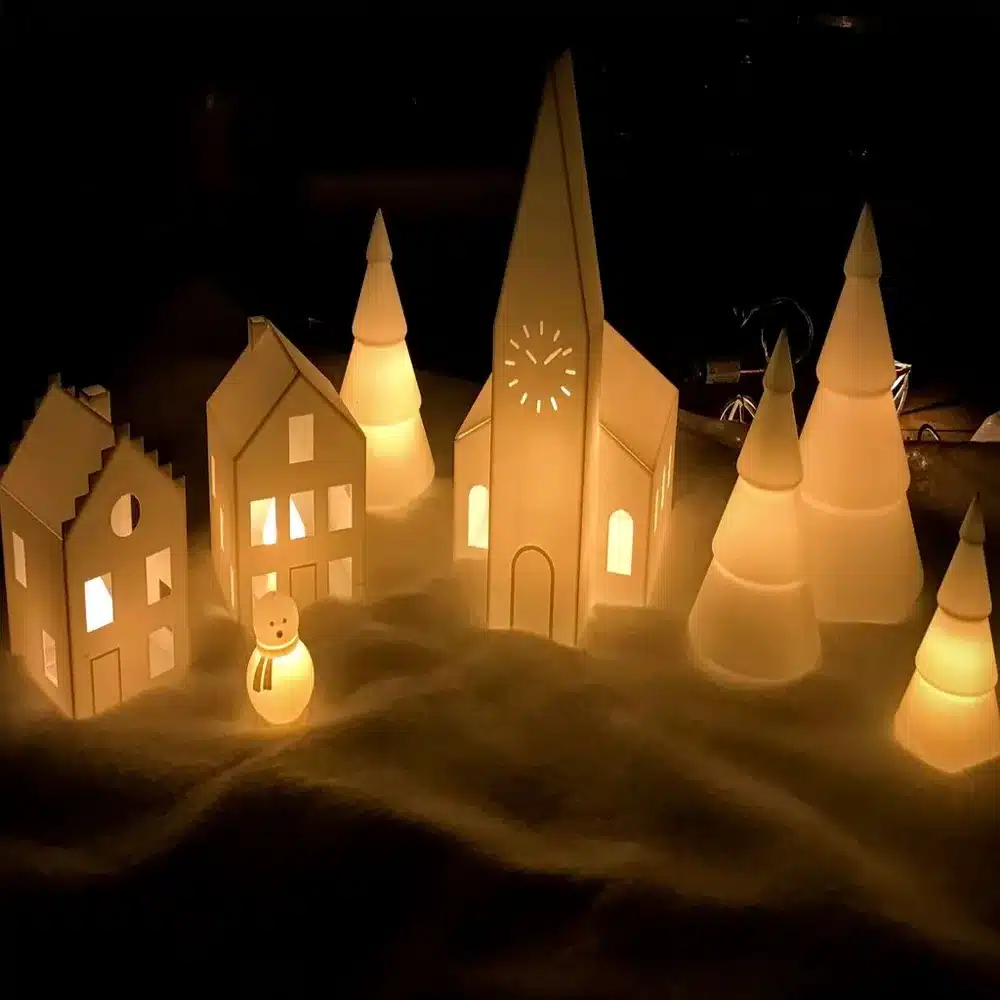 Cozy paper village scene with illuminated houses, church, trees, and snowman, evoking a warm, festive atmosphere at night.