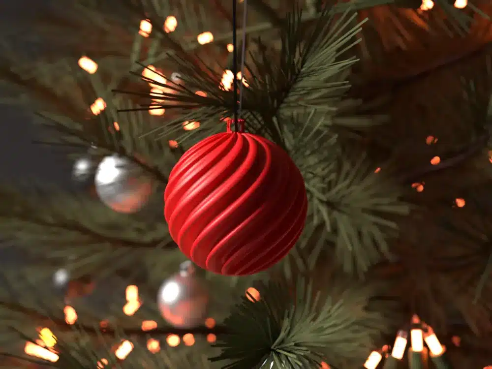 Red Christmas ornament hanging on a tree with festive lights and silver decorations in the background.