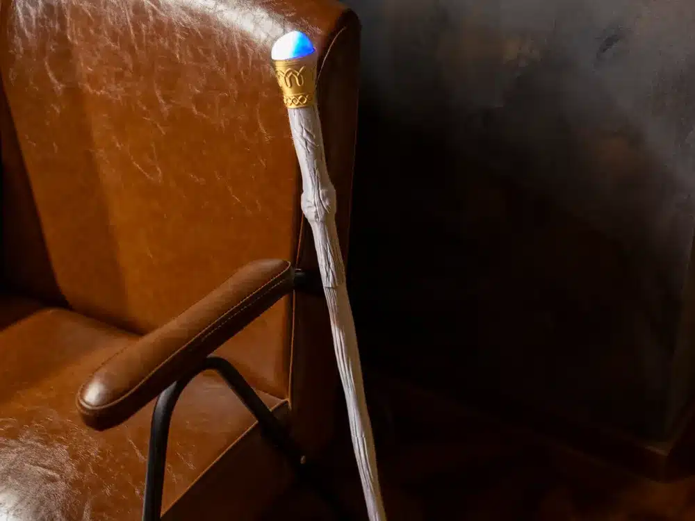 Illuminated wizard staff beside a leather chair in a dimly lit room, showcasing fantasy decor and cozy atmosphere.