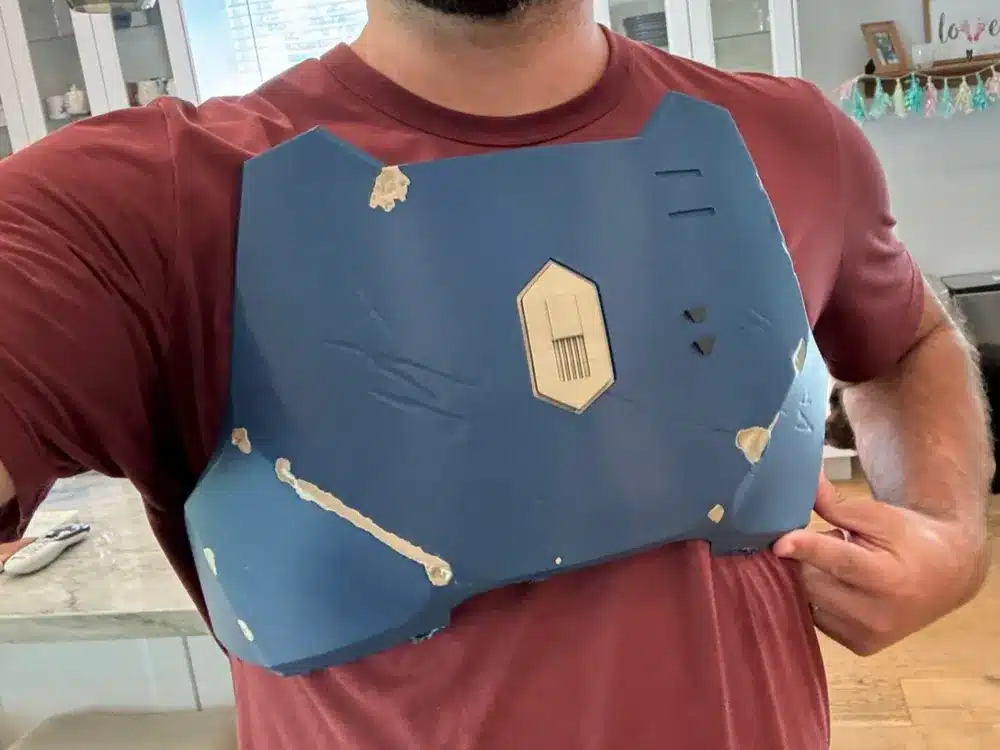 Man wearing homemade blue chest armor made from cardboard, standing indoors with a maroon shirt underneath.