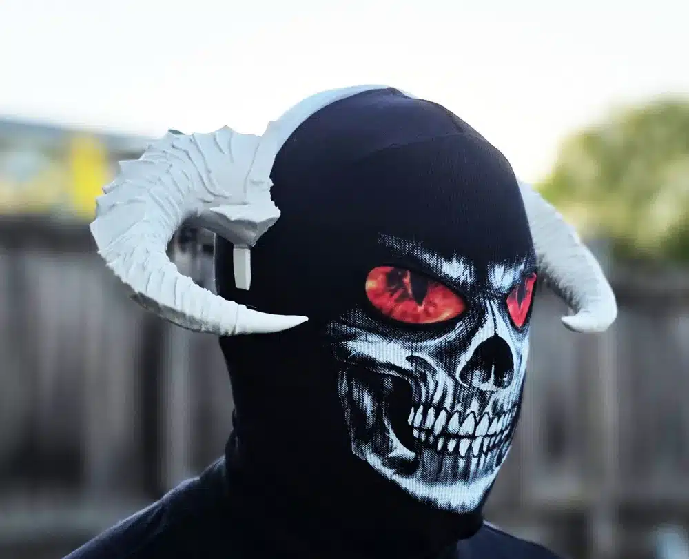 Man wearing a black mask with skull design and white horns, featuring glowing red eyes against a blurred background.