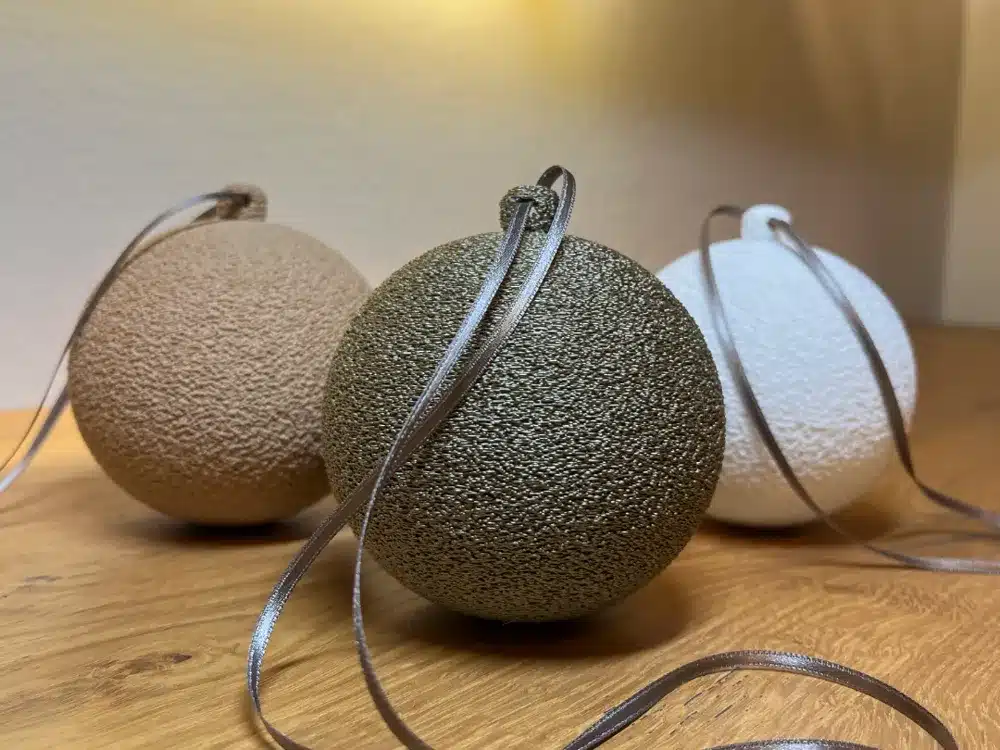 2025-10-27_b2053026f1d748 Three textured Christmas ornaments in brown, green, and white with ribbons on a wooden surface, lit by warm ambient light. | Sky Rye Design Three textured Christmas ornaments in brown, green, and white with ribbons on a wooden surface, lit by warm ambient light.