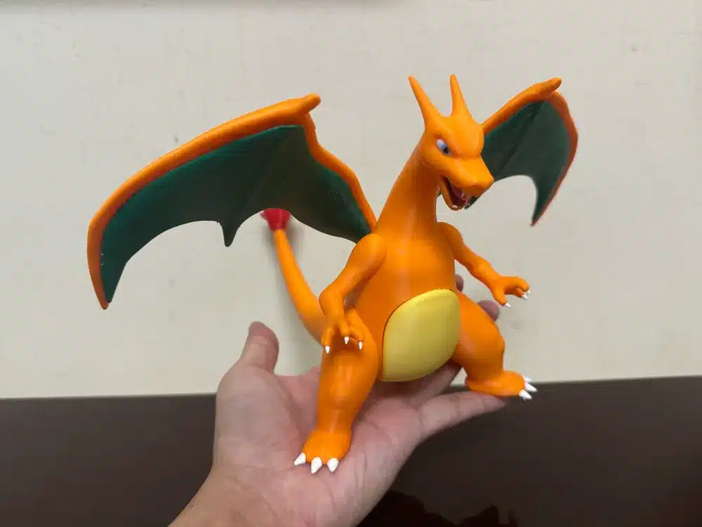 2025-10-31_e8961eba0b039 Hand holding Charizard figure with wings spread, orange body, and green wing accents. | Sky Rye Design Hand holding Charizard figure with wings spread, orange body, and green wing accents.