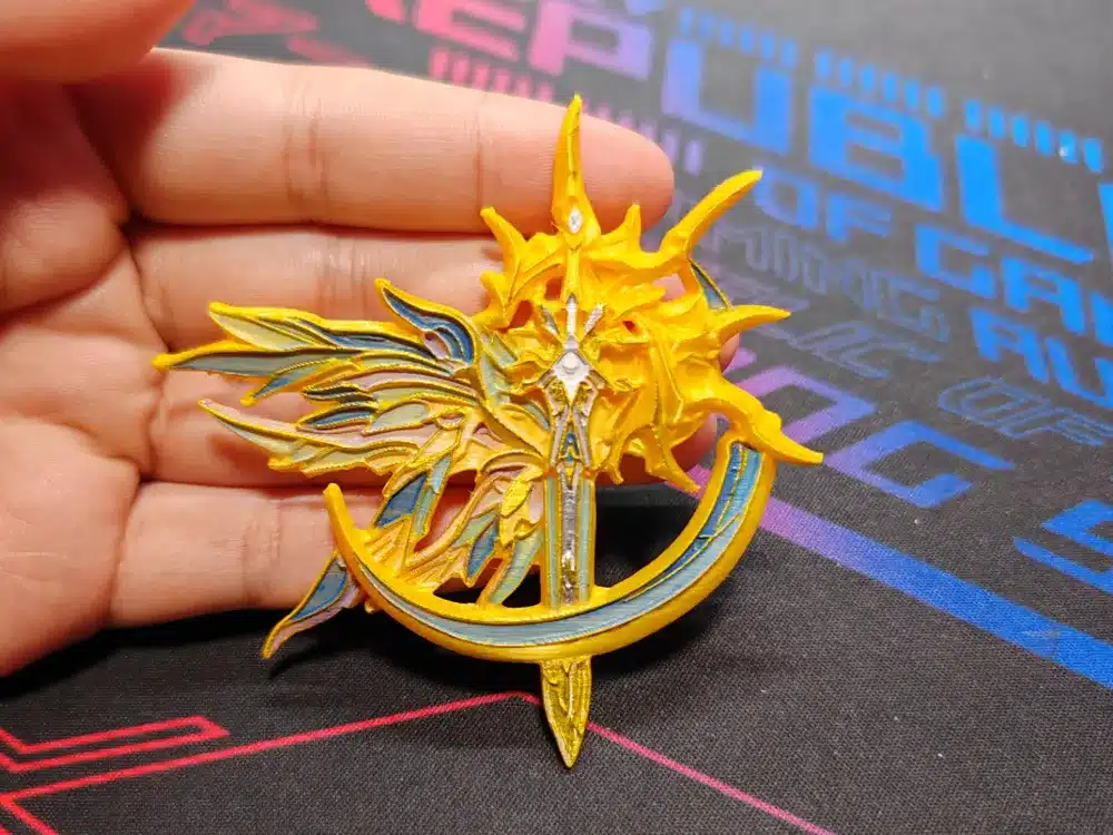 Intricate yellow and blue fantasy emblem with sun, moon, and wing motifs held in hand on a colorful gaming mat background.