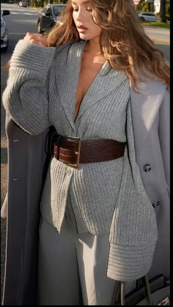 Fashionable woman in a cozy grey sweater and coat, styled with a wide brown belt, standing on a sunlit city street.