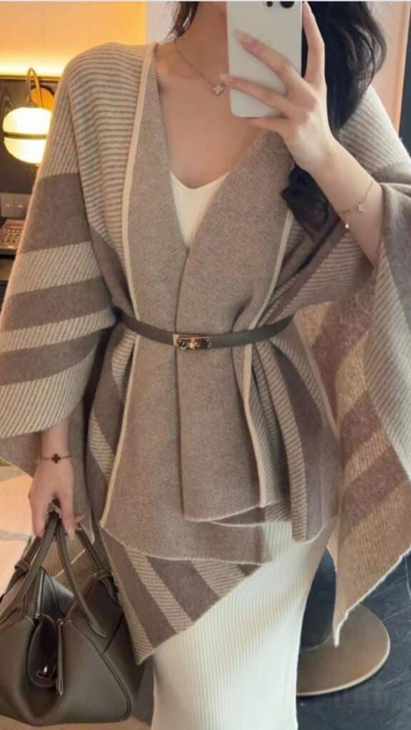 Woman in striped brown poncho and white dress taking a mirror selfie, holding a handbag, showcasing elegant fall fashion style.