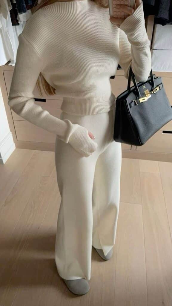 Woman in cozy white knit sweater and pants holding a black handbag, showcasing minimalist fashion in a modern closet setting.