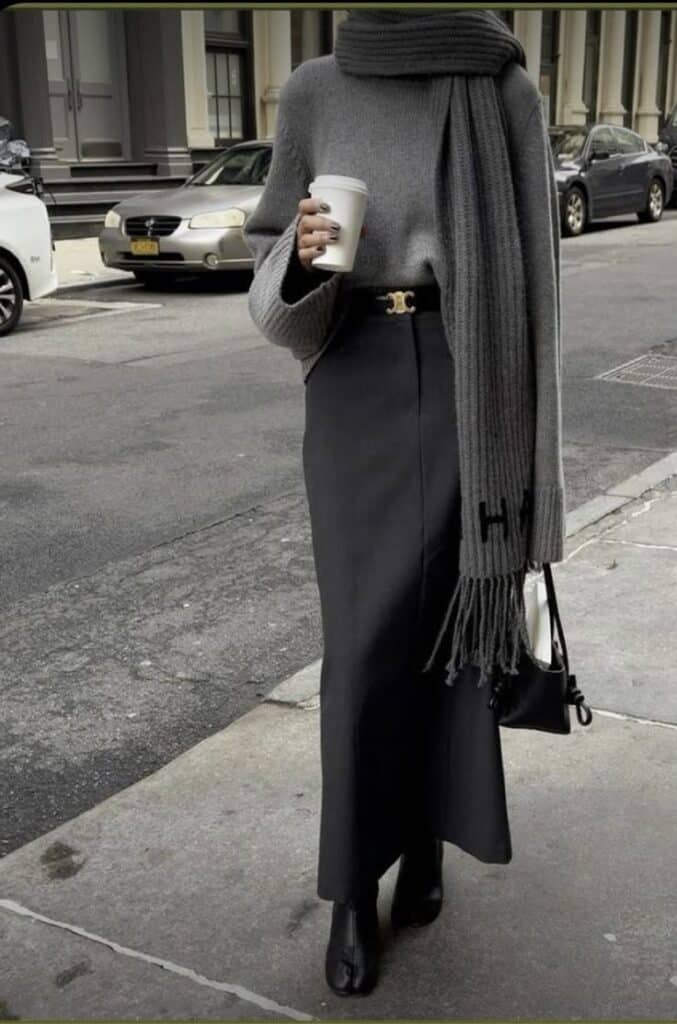 Stylish woman in a chic gray sweater, scarf, and black skirt, holding a coffee cup while walking on an urban street.