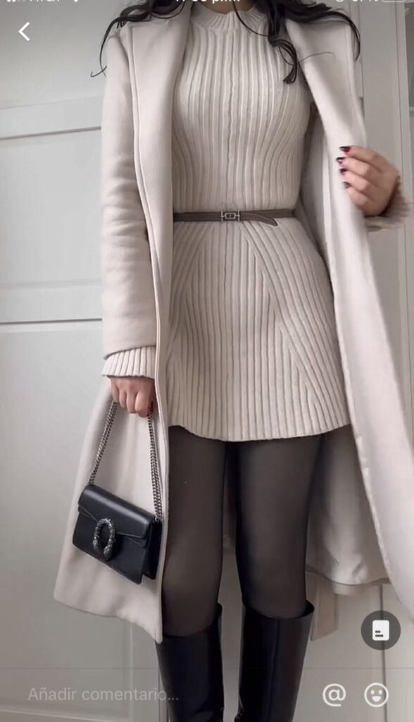 Elegant winter outfit featuring a beige dress, long coat, black tights, and boots, accessorized with a belt and chic handbag.