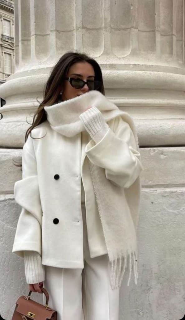 Woman in a chic white outfit with a scarf and sunglasses, standing by a column, holding a purse, showcasing stylish winter fashion.