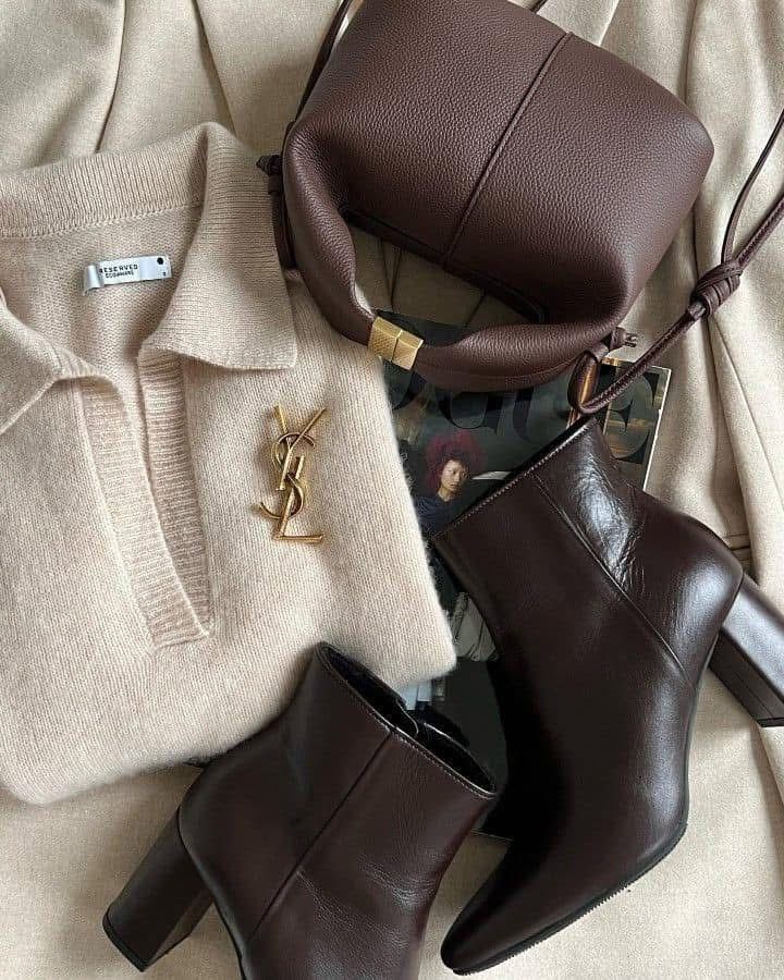 Beige sweater, designer logo, brown leather bag, and heeled boots on a stylish, monochrome backdrop with a fashion magazine.