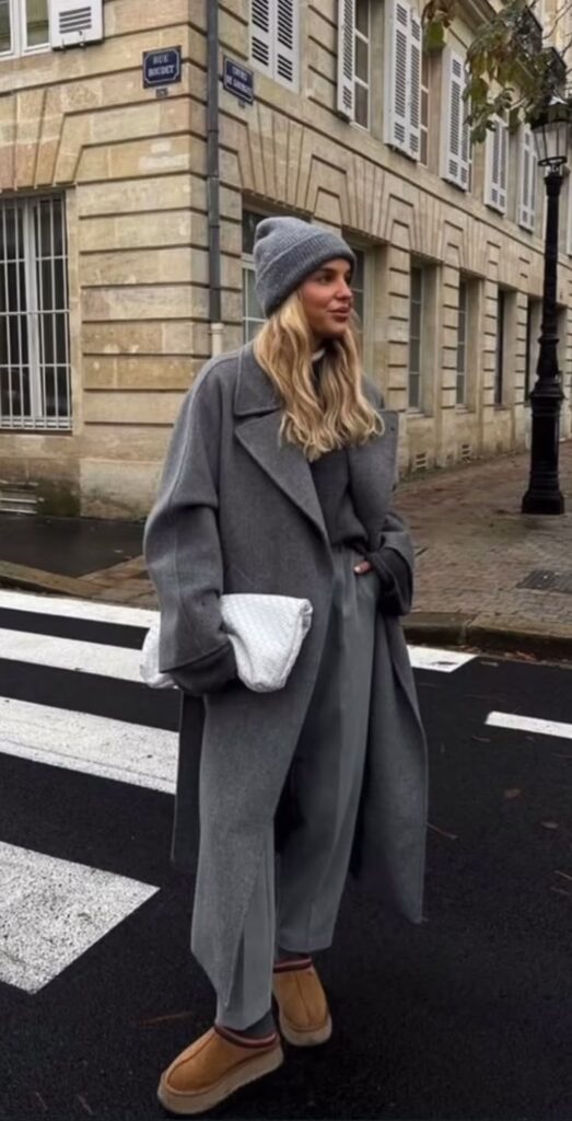 Woman in chic gray outfit with beanie and crossbody bag, standing on a Parisian street near historic buildings.