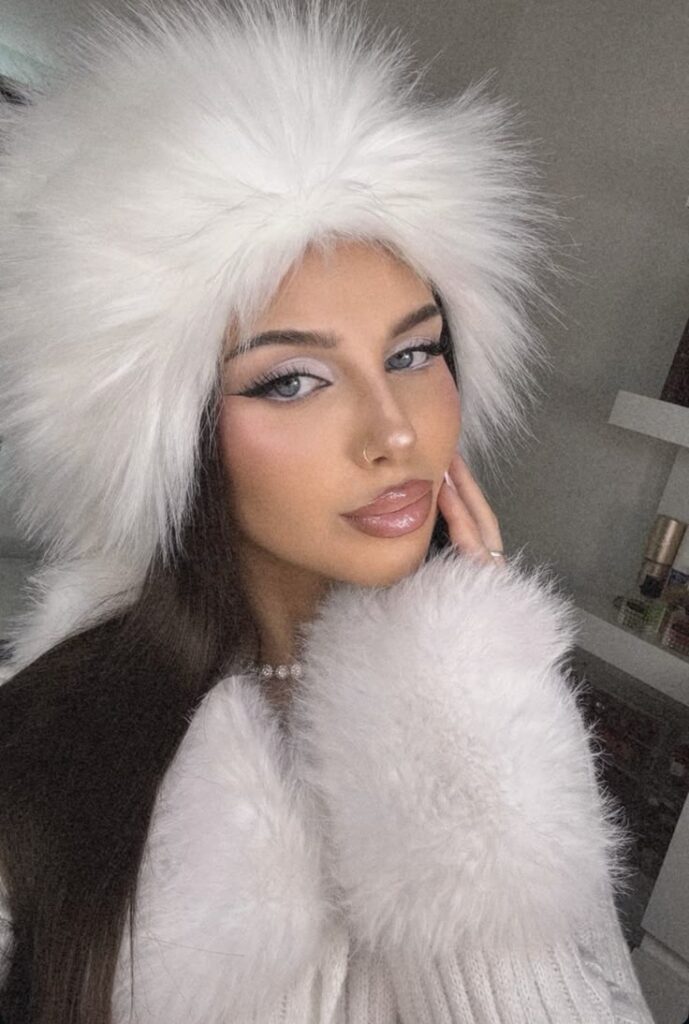 A woman wears a stylish white fur hat and coat, showcasing winter fashion. Her makeup is elegant and enhances her eyes, lips, and outfit.