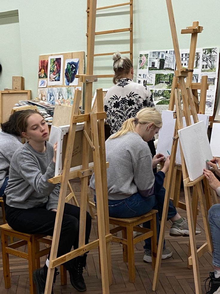 Art students drawing on easels in a classroom, surrounded by various paintings and sketches displayed on the walls.