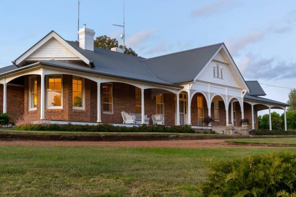 Charming brick house with white trim and wraparound porch surrounded by greenery at sunset, showcasing classic architectural design.