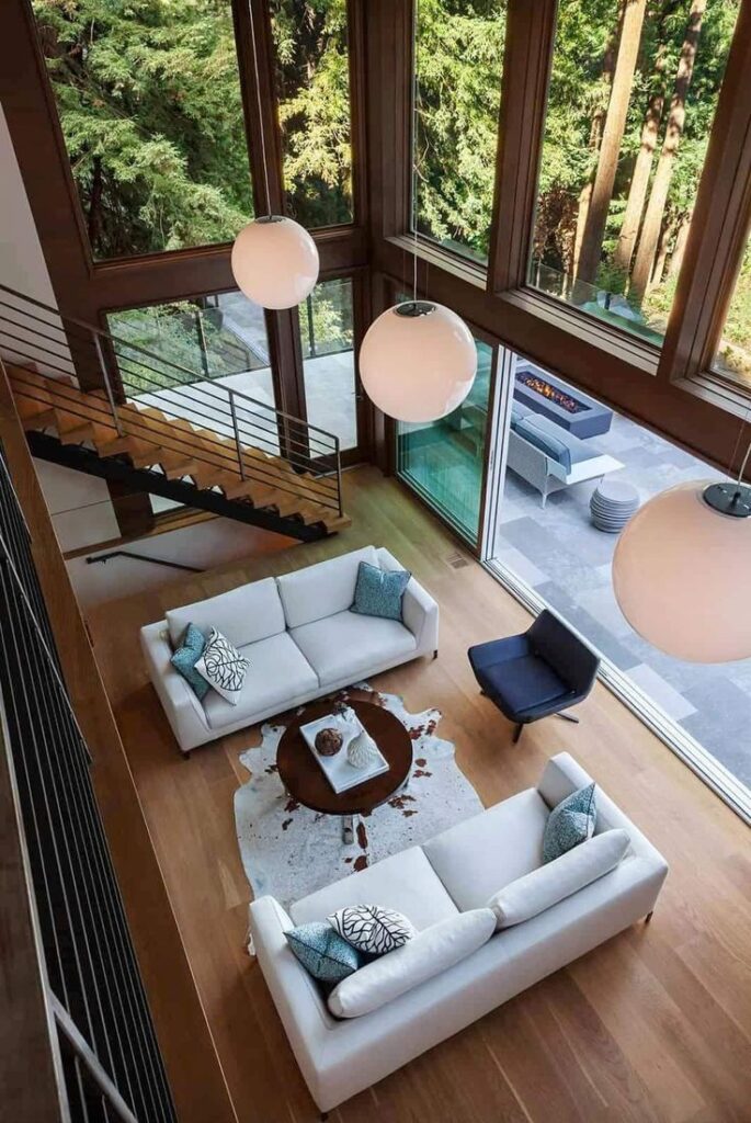 _ 25 Modern living room with large windows, white sofas, and forest view, featuring stylish pendant lighting and open staircase. | Sky Rye Design Modern living room with large windows, white sofas, and forest view, featuring stylish pendant lighting and open staircase.