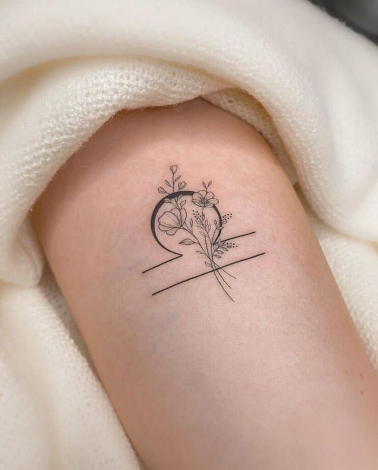 Delicate Libra tattoo featuring floral design on arm, surrounded by cozy white fabric, symbolizing balance and elegance.