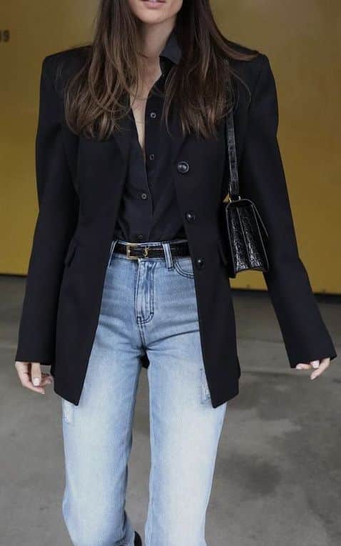_ 26 Fashionable woman in black blazer and jeans, accessorized with black belt and handbag, posing against a yellow background. | Sky Rye Design Fashionable woman in black blazer and jeans, accessorized with black belt and handbag, posing against a yellow background.