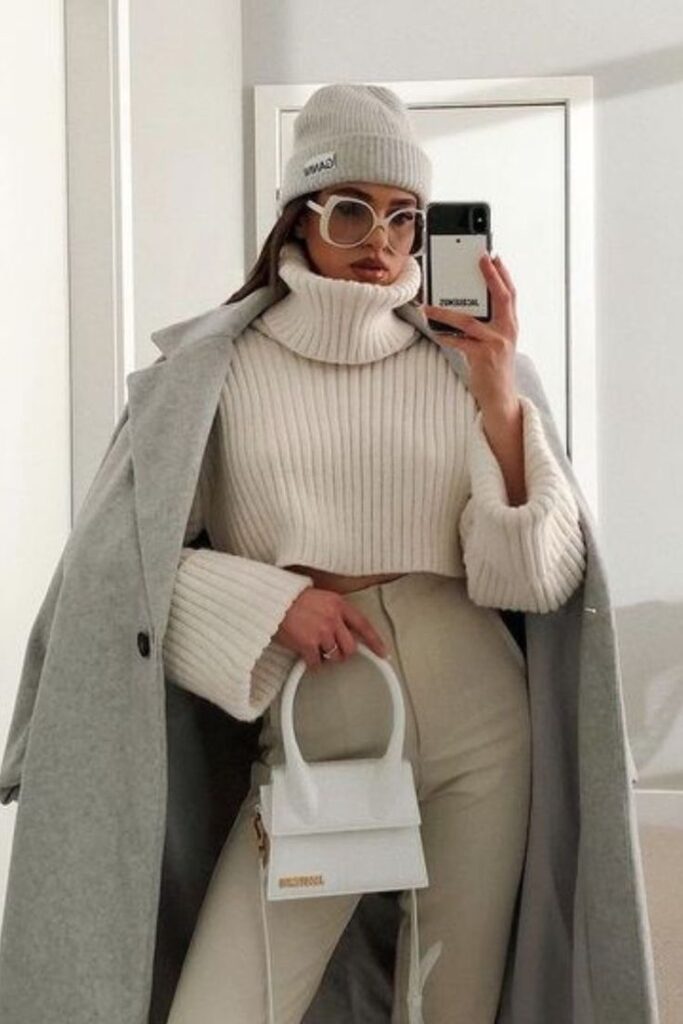 Fashionable woman in beige turtleneck and coat, mirror selfie, wearing beanie and glasses, holding a chic white handbag.