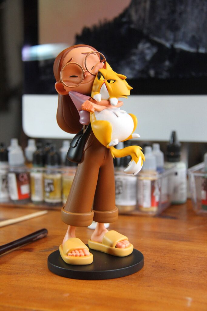 264fcee2a81c257b3fca9aa66a90a79e Cute cartoon figure with glasses hugging a grumpy yellow cat, on a desk with art supplies in the background. | Sky Rye Design Cute cartoon figure with glasses hugging a grumpy yellow cat, on a desk with art supplies in the background.