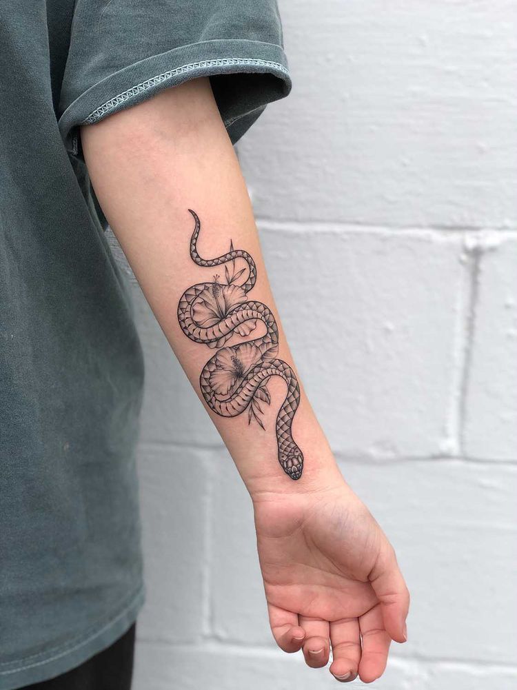 Snake tattoo on forearm featuring floral design, shown on a person's arm against a light gray brick wall background.