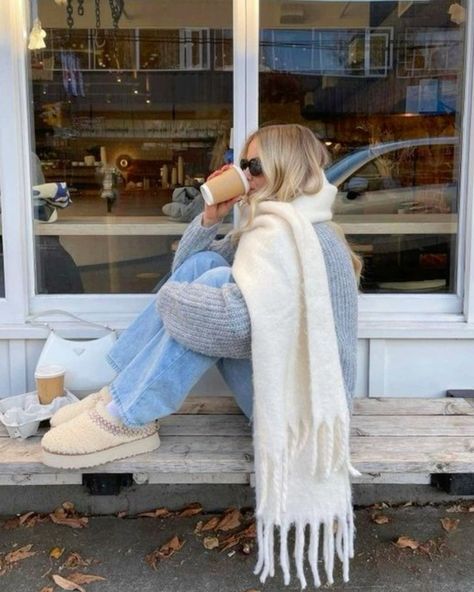 Woman in cozy outfit sipping coffee outside a café, wearing a white scarf, sunglasses, and beige shoes.
