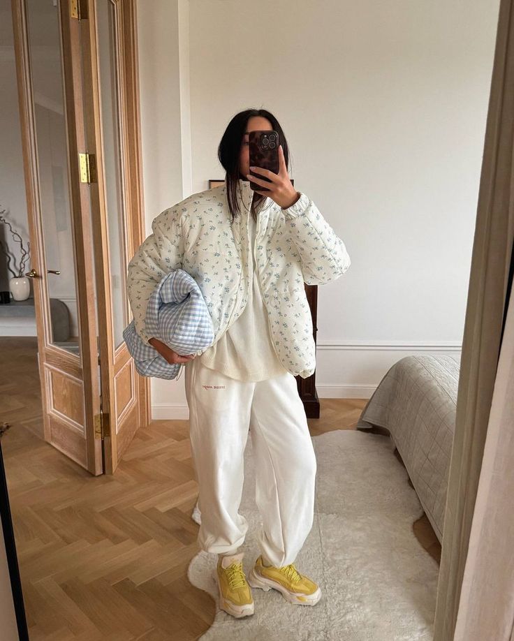 Person in cozy white outfit with patterned jacket and yellow sneakers, holding a blue-checkered pillow in a modern room. winter outfits