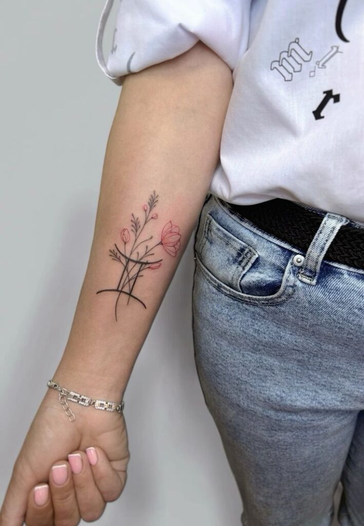 Minimalist floral tattoo on forearm, featuring delicate outlined flowers and subtle pink highlights, complementing casual jeans and white shirt.