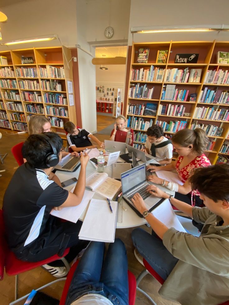 Students collaborating in a library, working on laptops and taking notes around a table. Bookshelves filled with books in the background.
