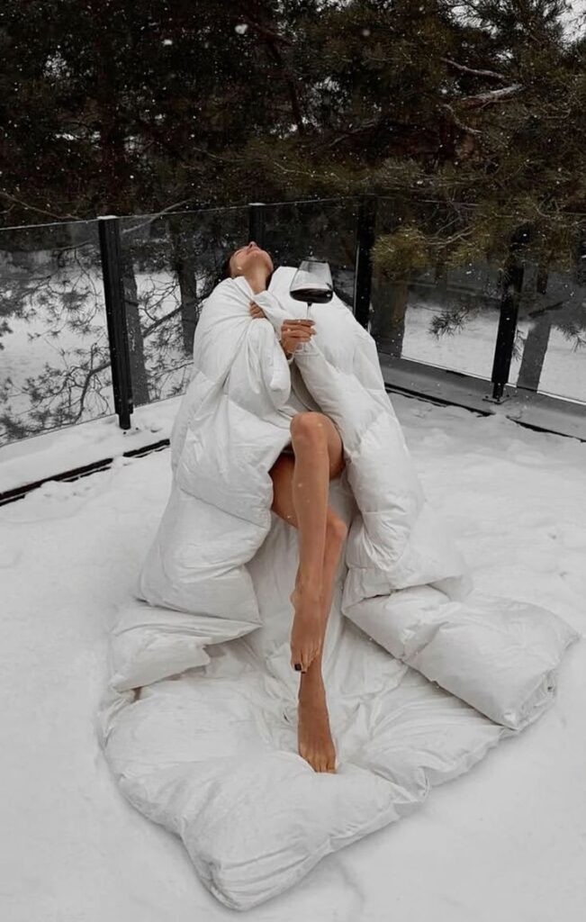 Person relaxing in snow with a comforter, holding a glass of wine, surrounded by trees.