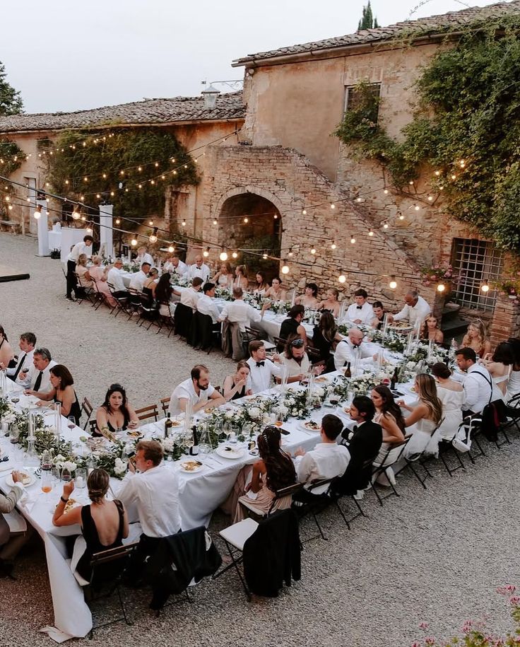 _ 3 Outdoor wedding reception with guests seated at long tables, strung lights, rustic stone building backdrop, elegant dinner setting. | Sky Rye Design Outdoor wedding reception with guests seated at long tables, strung lights, rustic stone building backdrop, elegant dinner setting.