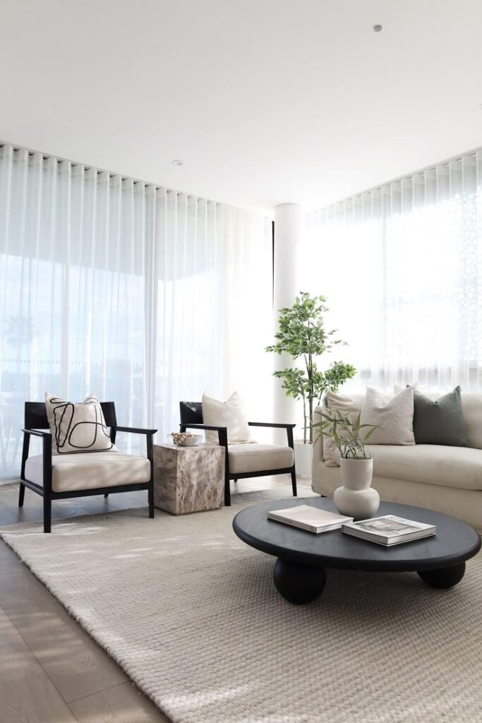Modern living room with cozy beige sofa, armchairs, potted plant, large rug, and a black round coffee table with books, flooded with sunlight.