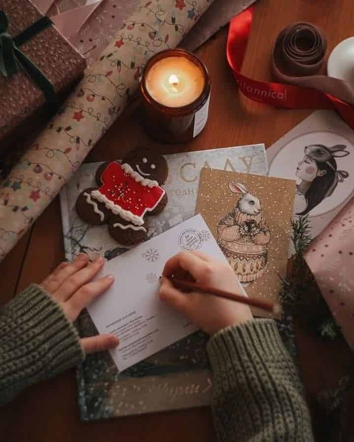 Person writing a holiday card surrounded by festive wrapping paper, candle, gingerbread cookie, and gift packaging on a wooden table.