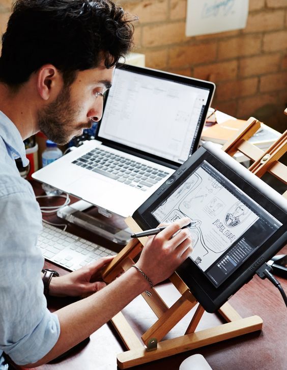 Designer working on digital art using a graphics tablet, with a laptop in the background, in a creative workspace.