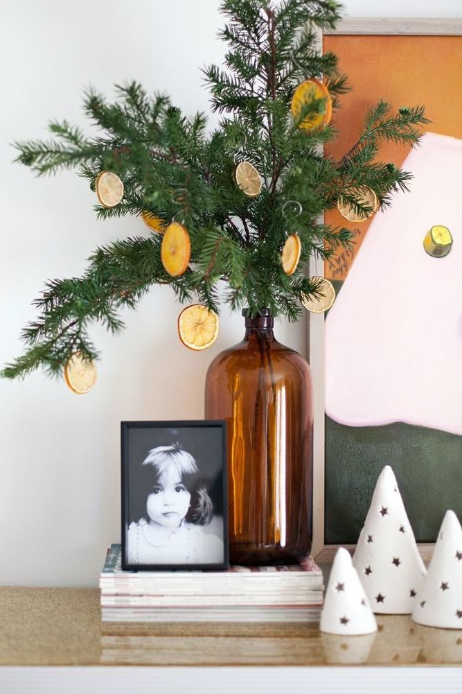 Decorative holiday scene with a pine branch in a brown vase, dried orange slices, a child's photo, and ceramic cone trees on a shelf.