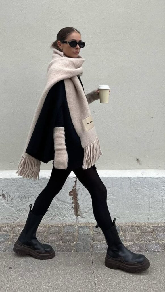 Stylish woman in oversized scarf, sunglasses, and boots walking with coffee against a neutral wall background.